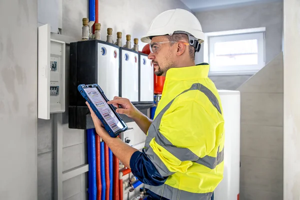 Man, an electrical technician in uniform working in a switchboard with fuses. Installation and connection of electrical equipment. Professional uses a tablet.