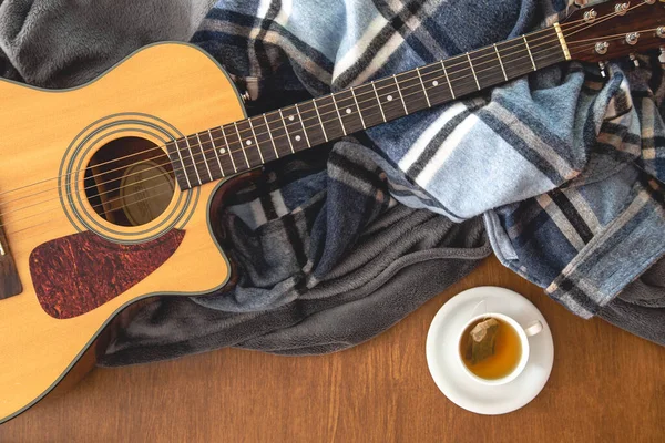 Acoustic guitar, plaids and a cup of tea on a wooden background, top view, the concept of musical creativity.