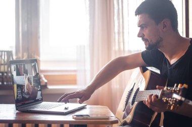 A man learning to play acoustic guitar, watching video lesson on laptop, concept of online learning and self-study.