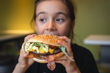 A little girl eats a big appetizing burger in a cafe, close-up.