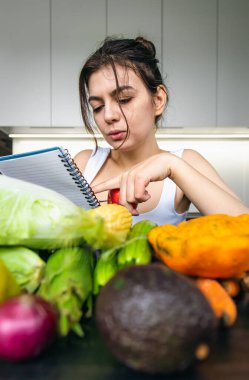 Mutfakta elinde sebzeler arasında not defteri olan genç bir kadın. Yemek pişirme, diyet yapma ve sağlıklı beslenme kavramı..