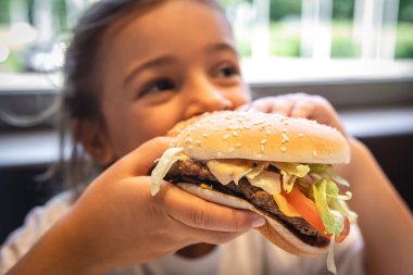 A little girl eats a big appetizing burger, close-up.
