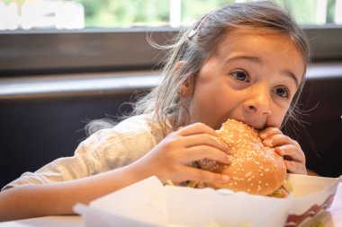 A little girl eats a big appetizing burger in a cafe, close-up.