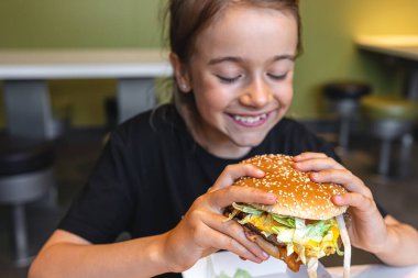 A little girl eats a big appetizing burger in a cafe, close-up.