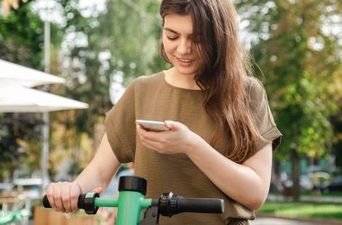 Attractive young woman with a smartphone in her hands rents an electric scooter on a sunny day.