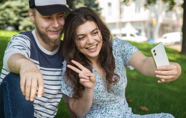Happy boyfriend and girlfriend take a selfie while sitting on the grass in the park.