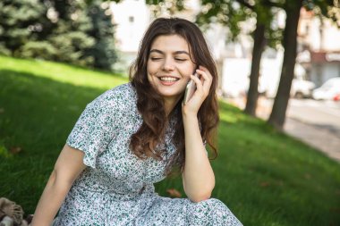 A young woman is talking on a smartphone while sitting on the grass in a park on a summer day.