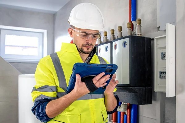 Man, an electrical technician in uniform working in a switchboard with fuses. Installation and connection of electrical equipment. Professional uses a tablet.