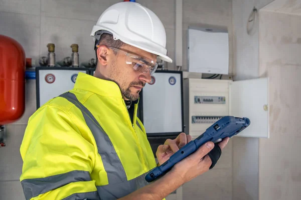 Man, an electrical technician in uniform working in a switchboard with fuses. Installation and connection of electrical equipment. Professional uses a tablet.