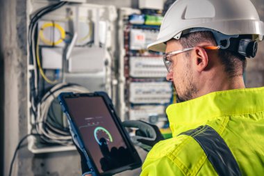 Elektrik santralinde fitillerle çalışan bir elektrik teknisyeni. Elektrik ekipmanlarının kurulumu ve bağlantısı. Profesyonel bir tablet kullanır.
