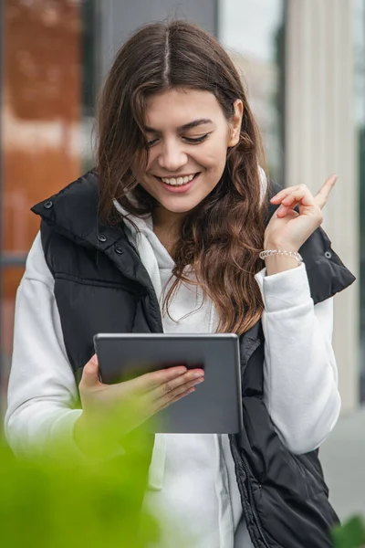 Attractive young woman stands outside and uses a tablet against a blurred background of a building.