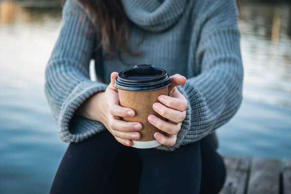 Close-up, a cup of coffee in the hands of a woman in nature by the river, a takeaway drink in the cold season.