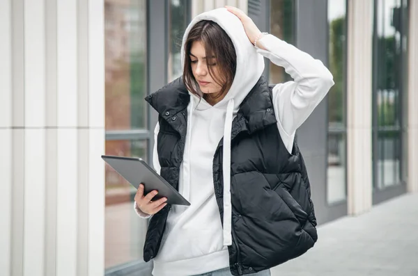 Business young woman stands outside and uses a tablet against a blurred background of a building.