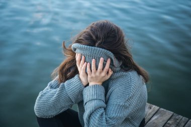 Bir kadın deniz kenarında otururken yüzünü bir kazağın içine sakladı. Ruh sağlığı ve ruh hali kavramı..