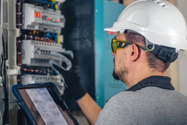Elektrik santralinde fitillerle çalışan bir elektrik teknisyeni. Elektrik ekipmanlarının kurulumu ve bağlantısı. Profesyonel bir tablet kullanır.