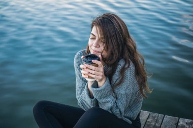 Bir fincan kahve içen genç bir kadın sabahın erken saatlerinde nehrin kenarında oturur ve doğanın tadını çıkarır..