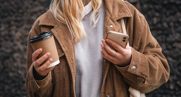 Close-up, a cup of coffee and a smartphone in the hands of a stylish woman in a corduroy jacket.