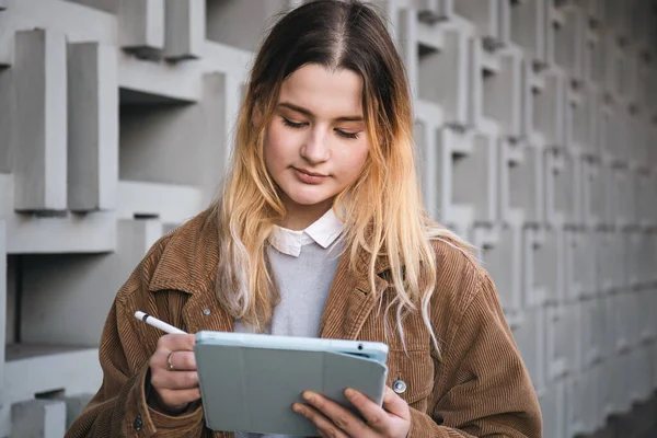 Attractive young woman in a corduroy jacket draws with a pencil on a digital tablet, on a blurred background of a geometric concrete wall in the city..