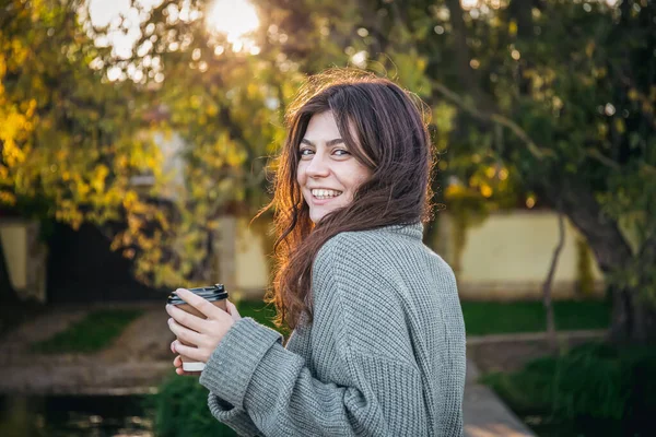 A young woman in a sweater with a cup of coffee in nature on a blurred background in the early autumn morning.