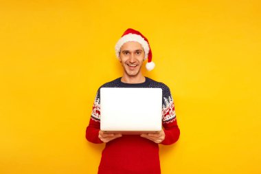 Noel Baba şapkalı, kırmızı Noel Baba şapkalı, gülümseyen, heyecanlı bir adam. İnternetteki dizüstü bilgisayarlı. Noel terfilerinde sevinçten havalara uçuyor.
