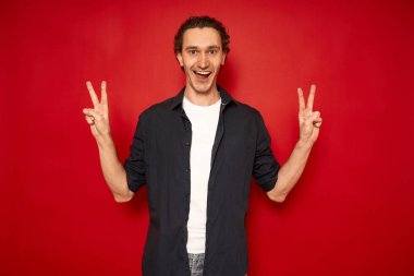 Heyecanlı, yakışıklı, güler yüzlü, sıradan elbiseli, neşeli adam mavi gömlekli, zafer işareti V 'yi elleriyle gösteriyor. Parlak kırmızı arkaplanda izole edilmiş, metin için boşluk var. konsept - kazanmak, barış, kutlama