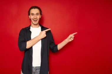 Genç, şaşkın, şaşkın, mavi tişörtlü erkek öğrencinin işaret parmağıyla boş işyerine gitmesi kırmızı arka plan stüdyo resminde izole edilmiş fotokopi alanının taklidini yapıyor. promosyon
