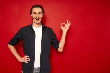 Gündelik giysiler içinde heyecanlı, mavi tişörtlü, beyaz tişörtlü genç yakışıklı adam, tamam işaretli kırmızı arka plan, mesaj, konsept - başarılı alışveriş, Noel indirimleri, promosyonlar