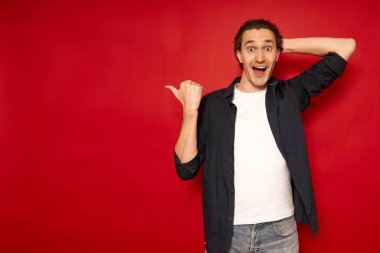 Genç, şaşkın, şaşkın, mavi tişörtlü erkek öğrencinin işaret parmağıyla boş işyerine gitmesi kırmızı arka plan stüdyo resminde izole edilmiş fotokopi alanının taklidini yapıyor. promosyon