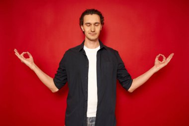 Gülümseyen, mavi gömlekli genç adam yoga jestinde el ele tutuşuyor, meditasyon yapıyor, sakin olmaya çalışıyor. Parlak kırmızı arka planda izole edilmiş, stüdyo resmi, fotokopi odası.