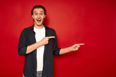 Genç, şaşkın, şaşkın, mavi tişörtlü erkek öğrencinin işaret parmağıyla boş işyerine gitmesi kırmızı arka plan stüdyo resminde izole edilmiş fotokopi alanının taklidini yapıyor. promosyon