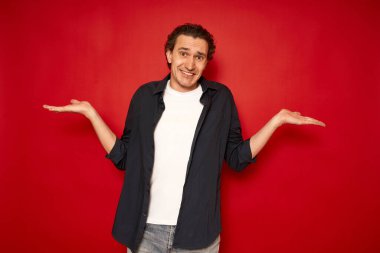 Çekici bir adamın yatay görüntüsü, kafası karışmış bir ifadeyle elleri açılır. Gülünç bir şekilde gülümser. Günlük kıyafetler giyer ve kırmızı arka planda izole edilmiş kararsız bir kızgınlık hisseder. Şüphe kavramı