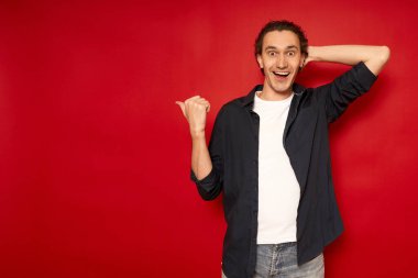 Genç, şaşkın, şaşkın, mavi tişörtlü erkek öğrencinin işaret parmağıyla boş işyerine gitmesi kırmızı arka plan stüdyo resminde izole edilmiş fotokopi alanının taklidini yapıyor. promosyon