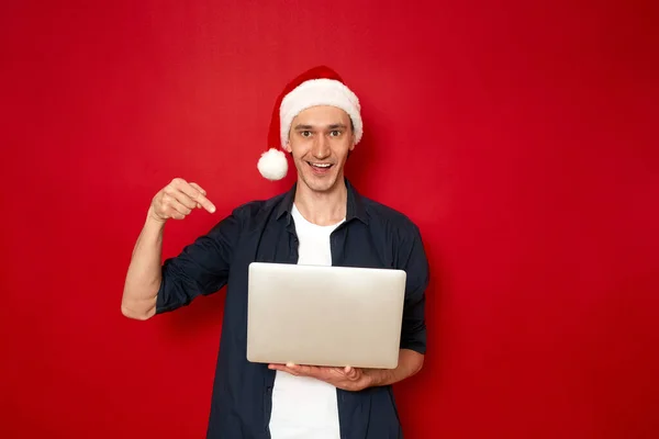 An excited smiling man in Santa Claus Christmas hat points with his index finger laptop in his hands recommends online purchases, sales, promotions, discounts isolated on red background space for text — Stock Photo, Image