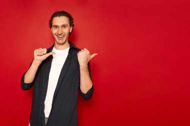 Yakışıklı, gülümseyen, neşeli bir adamın stüdyo portresi arkasında boş bir alana baş parmak basar, günlük kıyafetler giyer, kırmızı arka planda izole edilmiş mavi bir gömlek giyer. konsept - insanlar, tavsiye, onay