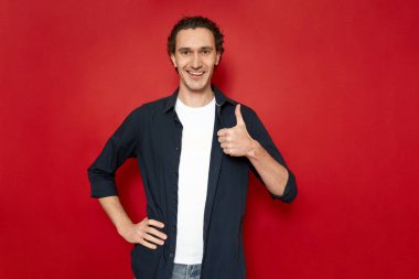 Yakışıklı, güler yüzlü, neşeli bir adam, kırmızı arka planda işaretlenmiş, günlük kıyafetler içinde mavi tişört ve yazı için boş alan olan başparmaklarını kaldırır. konsept - insanlar, öneriler, onaylar