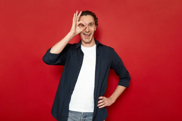 Agitated man brings his hand to his eye showing ok sign. dressed in casual clothes. isolated on red background with space for advertising text. concept - people, body language, luck, victory, planning — Stock fotografie