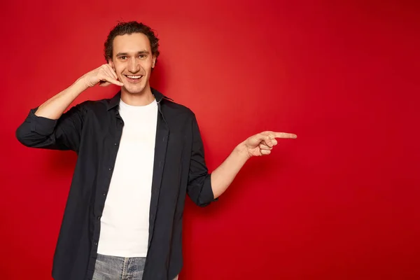 call me. man depicts telephone conversation hand, holding it near his ear, pointing with his index finger at an empty space for advertising text. dressed in casual clothes. isolated on red background.