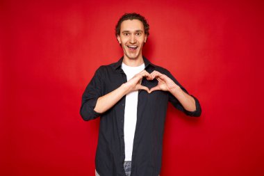 Neşeli, heyecanlı bir adam kalp işaretini göğsünün önünde gösterir ve aşkını ağzı açık itiraf eder. Kırmızı arkaplan kopyalama boşluğunda izole edildi. konsept - romantizm, ilişkiler, sevgililer günü