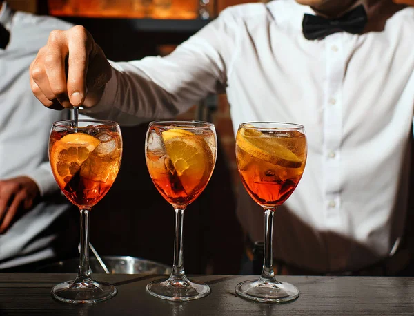 Three glass glasses with an aperol spritz cocktail are on bar. bartender in white shirt, bow tie is stirring one of aperitifs. concept - people, drinks, entertainment, recreation. free space for text. — Stock Photo, Image