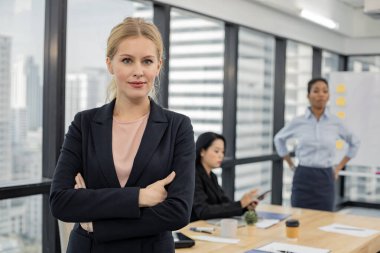 Diğer işçiler arka planda toplantı yaparken kollarını kavuşturmuş ve kendine güvenen güzel bir kadın. Toplantı odasında kameraya bakan beyaz iş kadınının portresi..