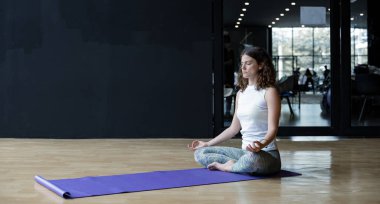 Beyaz kadın meditasyonda oturuyor, yoga minderinde yoga yapıyor spor salonundaki egzersiz odasında fotokopi odasıyla. Yoga yaparak egzersiz yapan bir kadın yoga konseptiyle evde yoga yapıyor..