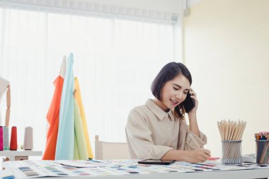 Güzel Asyalı kadın tasarımcı, müşteriyle telefonda konuşuyor ve müşteriye gülümseyerek terzi siparişini almak için iş detaylarını not alıyor. Başlangıç tasarımcısı konsepti.