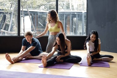 Group of young people doing yoga on a yoga mat with a trainer gradually teaching in exercise room. Young women, man and their teacher practicing yoga in gym. Concept of exercise by yoga. clipart