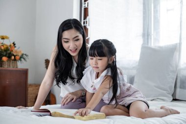 Mother is telling stories to her daughter on the white bed in the bedroom. Asian mum and daughter do fun activities at the bedroom. Happy loving family concept. Asian family.