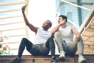 Koşucu Afrikalı genç adam ve beyaz adam spor kıyafetleri içinde selfie çekiyor, kameraya bakıyor ve dışarıda antremandan sonra merdivenlerde gülümsüyor..