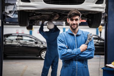 İş arkadaşı arka planda çalışırken kollarını kavuşturup gülümseyen üniformalı yakışıklı tamirci. Otomotiv servisinde çalışan araba tamircisi..