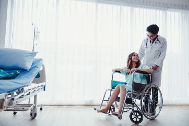 Woman patient in green shirt sitting on hospital wheelchair in patient's room with physician's care for the sick in hospitals closely. Healthcare and medical concept.