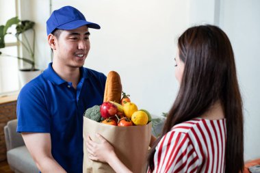 Service food delivery staff in blue uniform delivered fresh food to young woman customer at the home after receiving order from courier. Online shopping, Express food delivery service concept.