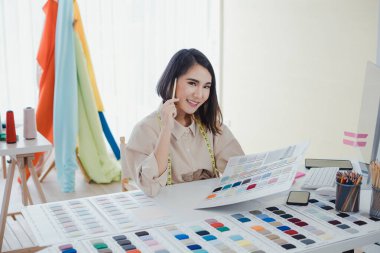 Asyalı kadın tasarımcı, stüdyodaki tasarımcı masasında müşteriler için kıyafet tasarlıyor ve düşünüyor. Kıyafet tasarımcıları ofiste çalışıyor. Başlangıç tasarımcısı konsepti.