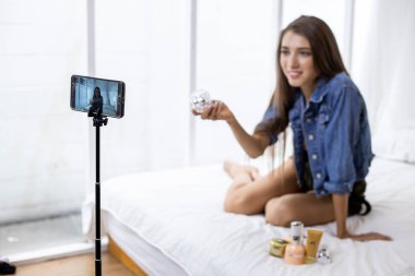 Young woman reviews and selling cosmetics online via her mobile phone on the bed at home. Sell stuff online. Live selling.